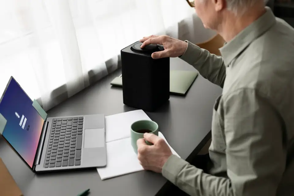 Ein älterer Mann arbeitet am Schreibtisch mit Laptop, Notizbuch und Kaffeetasse, während er einen smarten Lautsprecher bedient – Symbol für moderne Arbeitsprozesse mit CompanyGPT von snutig.