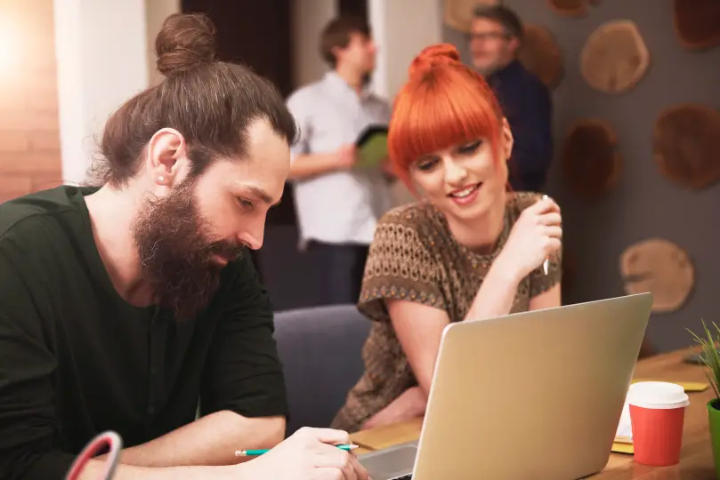 Zwei junge Menschen arbeiten gemeinsam an einem Laptop und besprechen Ideen, während im Hintergrund weitere Personen im Gespräch sind – kreative Zusammenarbeit bei Snutig